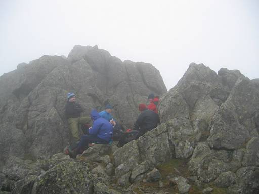 12_53-1.jpg - Very strong winds along the ridge have made going difficult. The lunch stop had to be made behind this rock and was still very chilly. Notice that Chris has a hat on.